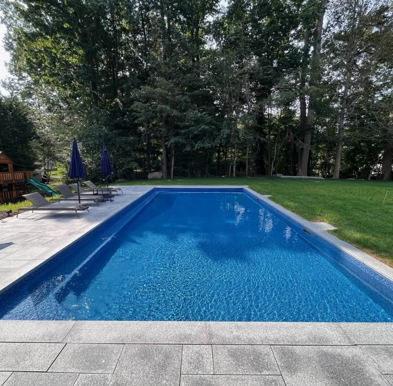 Rectangular in-ground pool with bright blue vinyl liner, light stone deck, and lounge chairs beside a wooded yard.