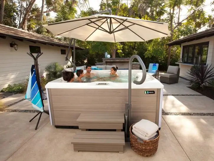 A family of four sits inside a tan-colored rectangular hot tub on a stone patio. A large white patio umbrella provides shade over the tub. A black towel rack with a blue striped towel and a wicker basket filled with white towels are next to the hot tub steps.