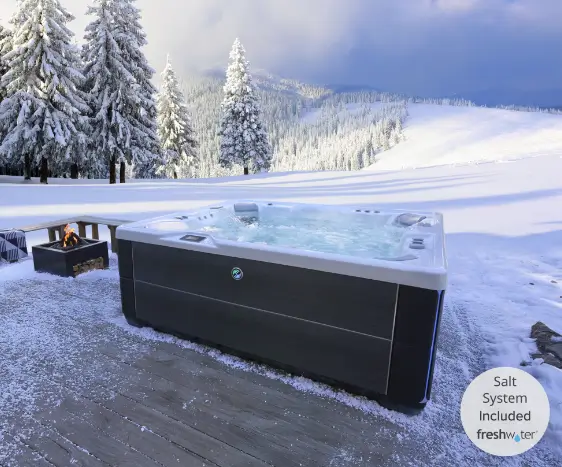 A dark grey hot tub with bubbling water sits on a snow-covered wooden deck. In the background, a pristine white snowy field leads to a forest of snow-laden evergreen trees under a cloudy sky. A small, square black fire pit with a flickering fire is visible to the left.