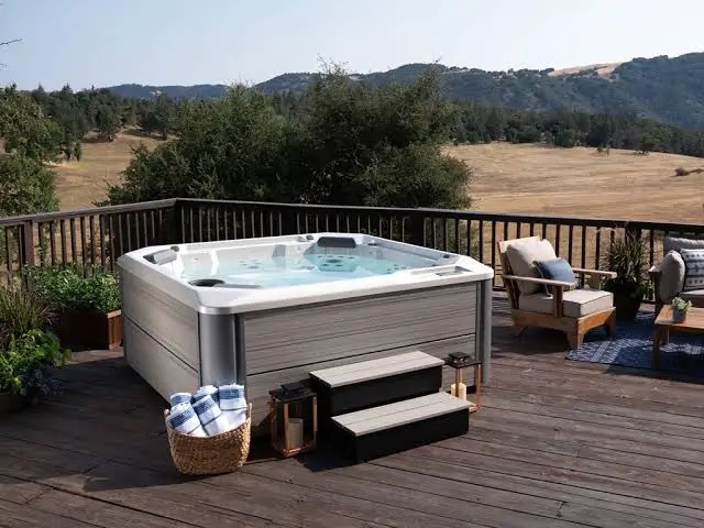 A grey rectangular hot tub is situated on a dark wood deck that overlooks a vast landscape of rolling hills and trees. On the deck, there's a wicker basket of blue and white striped towels, several black lanterns, and a wooden armchair with light-colored cushions.