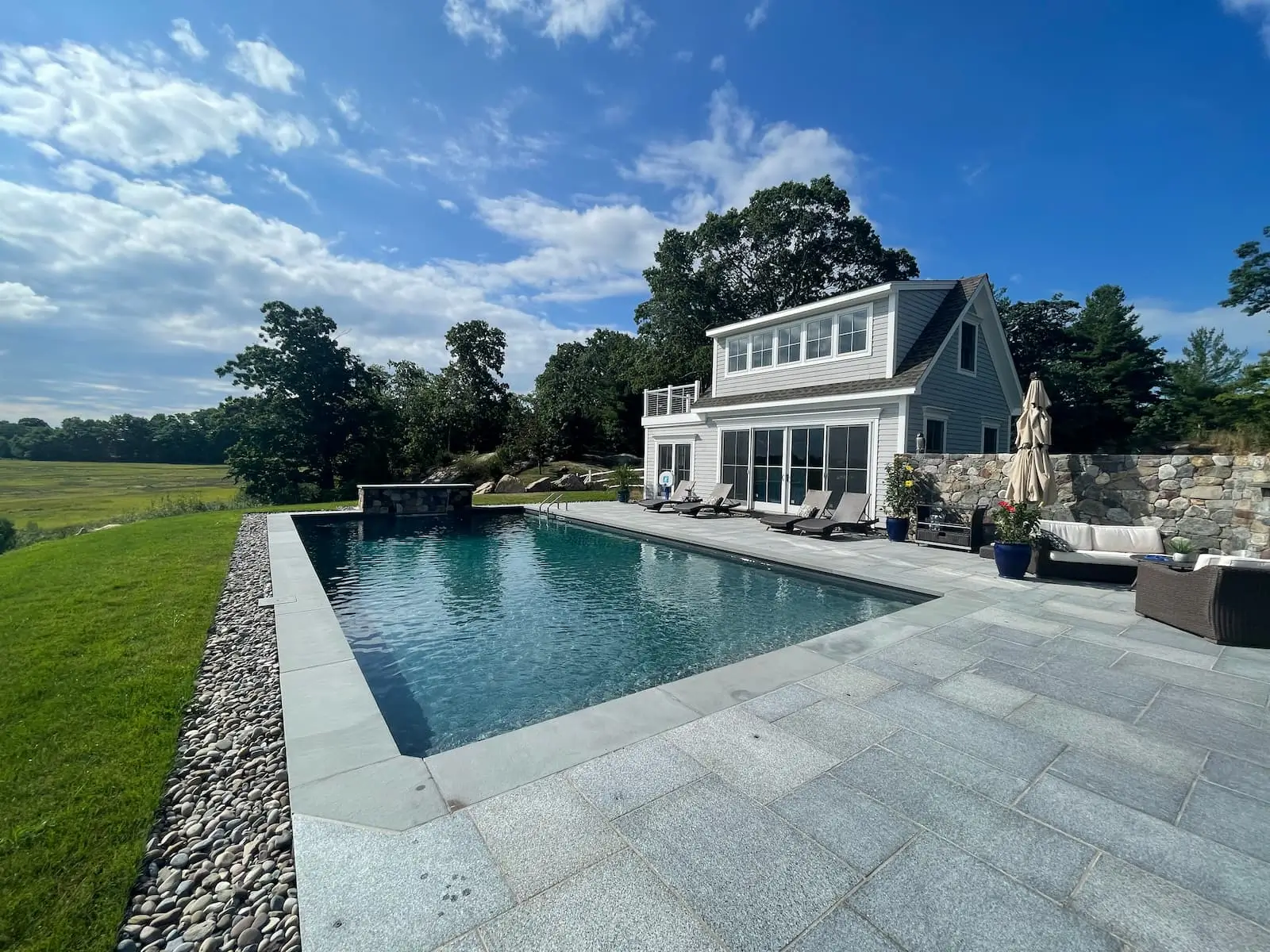 Rectangular pool with stone patio and lounge chairs next to a gray house under a blue sky.