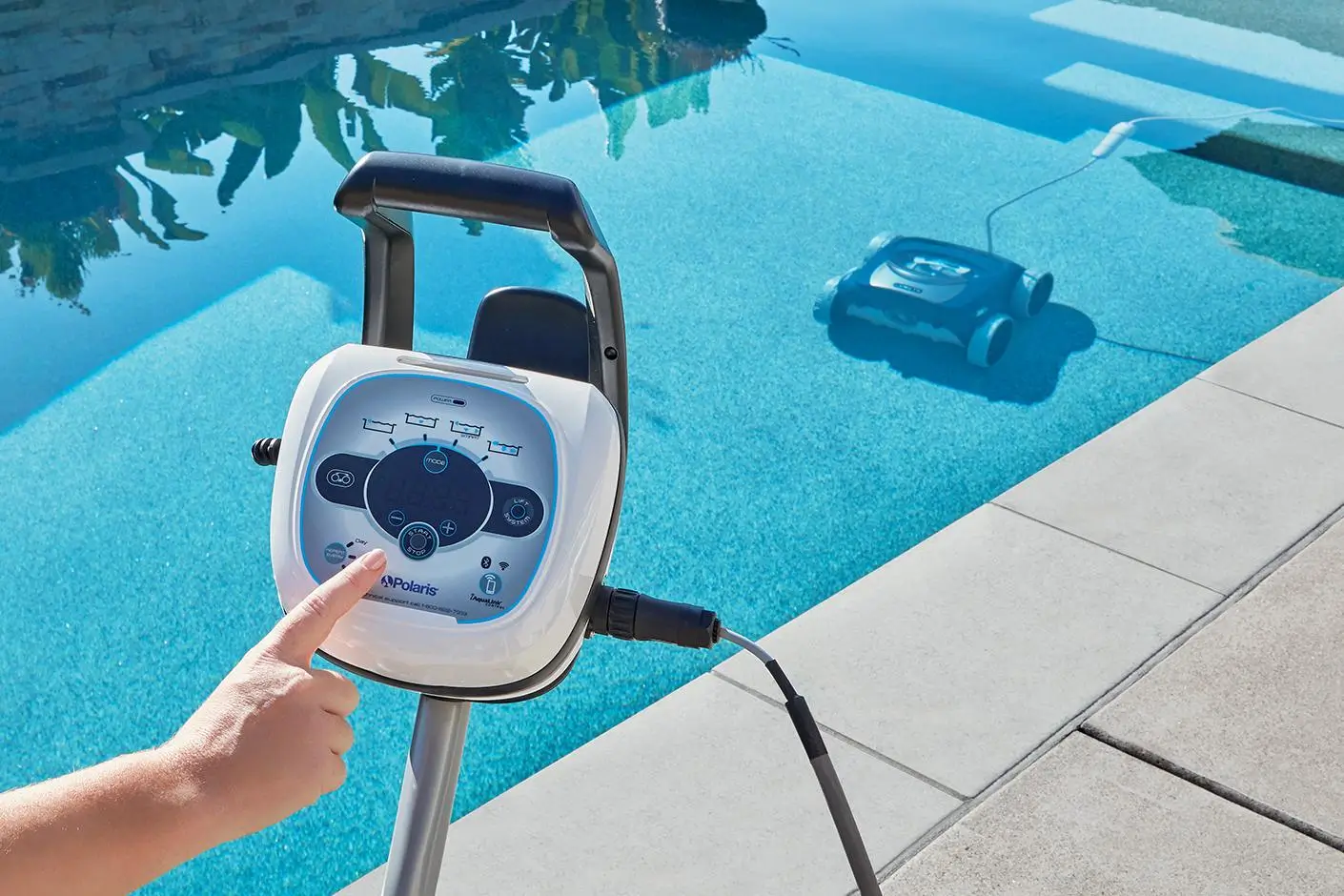 A close-up of a person's finger pressing a button on a white and blue Polaris control unit mounted on a stand next to a swimming pool. A robotic pool cleaner can be seen in the distance at the bottom of the pool.