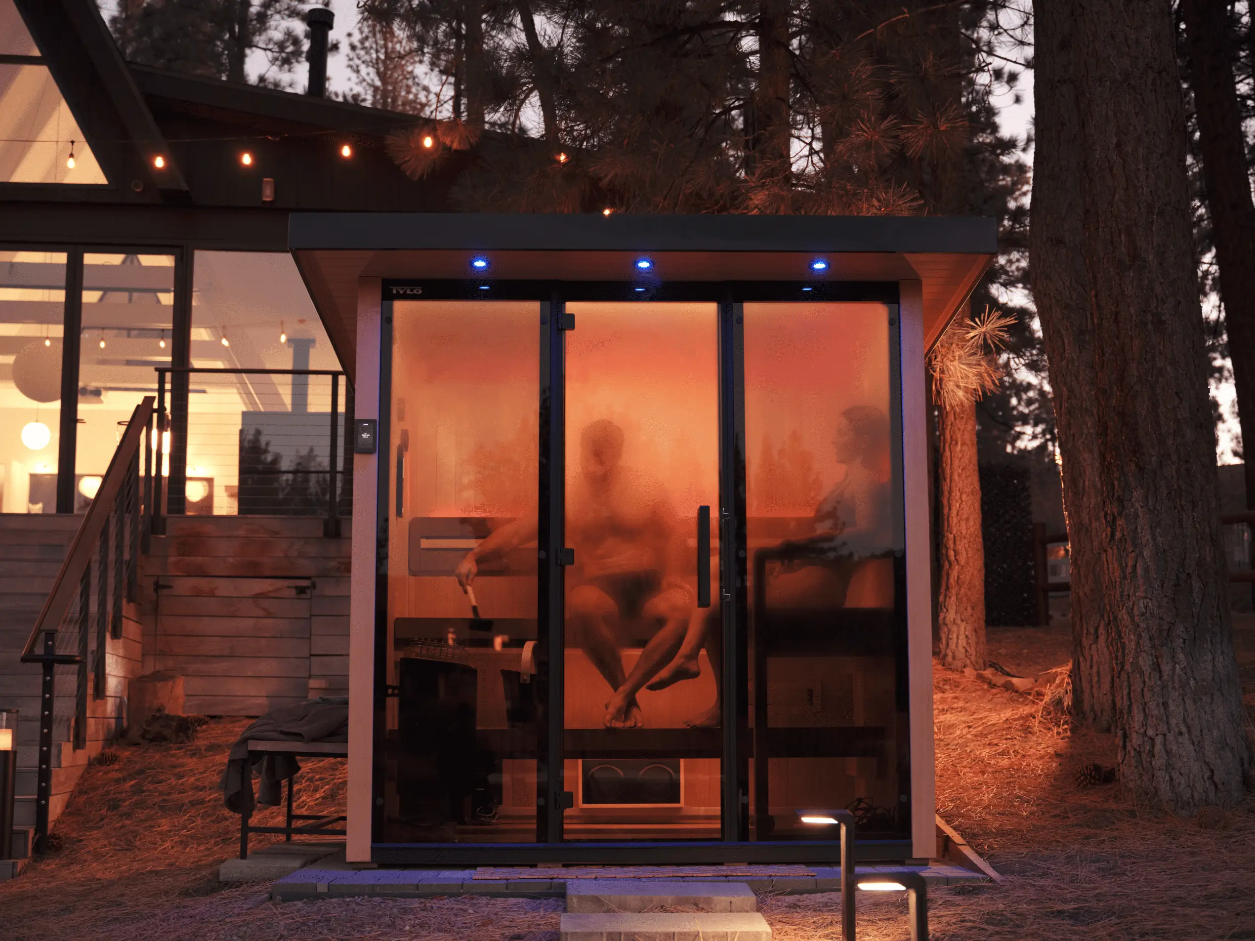 An outdoor Tylö sauna glows warmly at night with two people inside, situated in a wooded area near a cabin decorated with string lights.