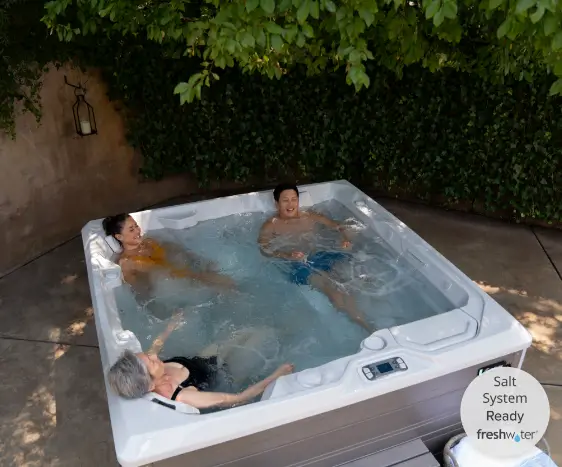 An overhead view shows three people – an older woman, a younger woman, and a man – relaxing in a square hot tub with clear, bubbling water. The hot tub is located in an outdoor area with a concrete floor and a backdrop of a stone wall covered in lush green ivy.