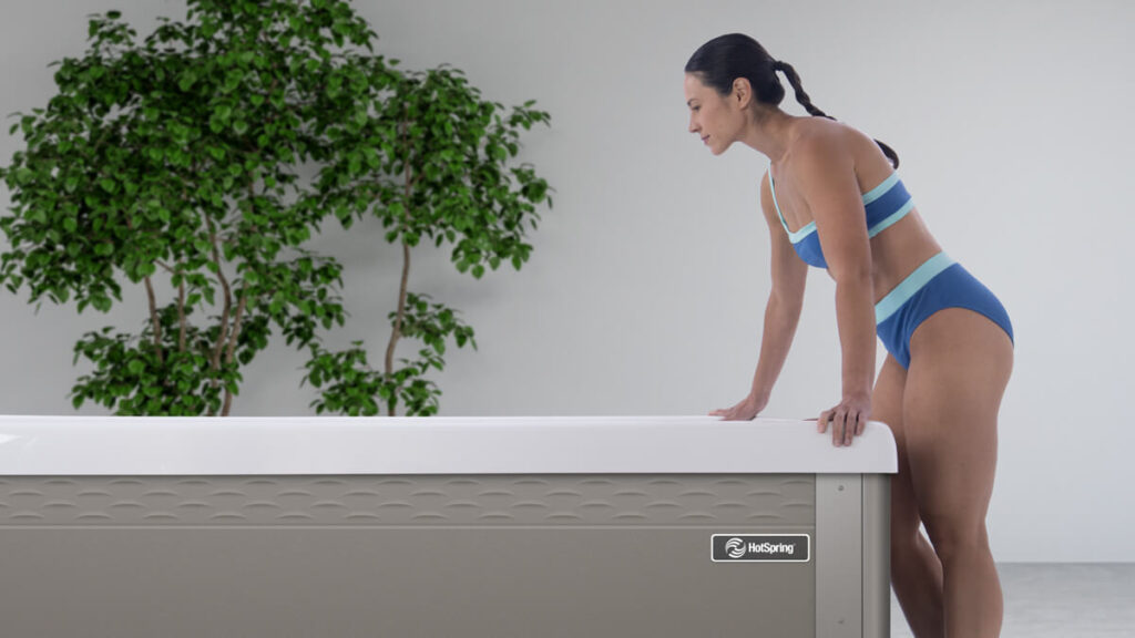 A woman in a blue and light-blue bikini leans over the side of a sleek, grey HotSpring cold plunge tub, preparing to step inside. A large green indoor plant stands in the background against a clean white wall.